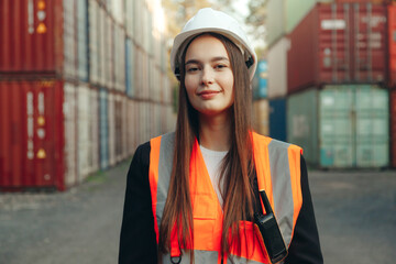 Portrait of beautiful engineer woman with walkie-talkie in white helmet and vest working in container terminal port