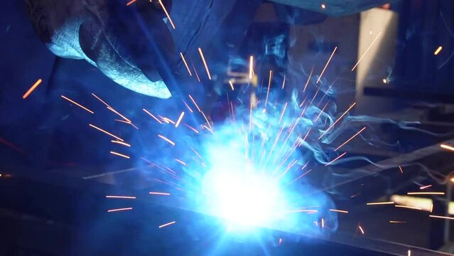Sparks fly from the hot metal. Slow motion close up.
  A working welder welds metal in a protective helmet, a beautiful epic shot, a work rack.