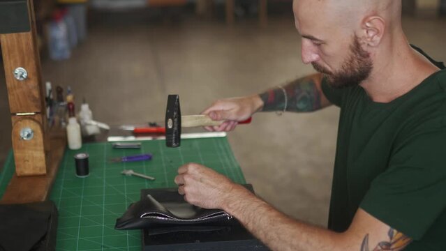 Side shot of professional leather craftsman at work table making stitching holes in black handbag with leather punch and hammer. Bold man with beard in khaki T-shirt sewing purse in workshop.
