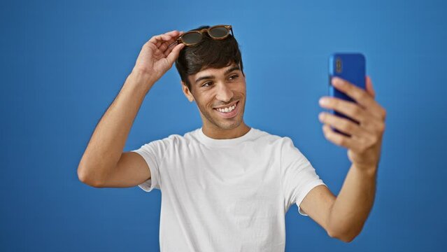 Cool-looking young hispanic man confidently donning sunglasses, capturing a fun selfie with joyous smile, standing isolated against blue wall.