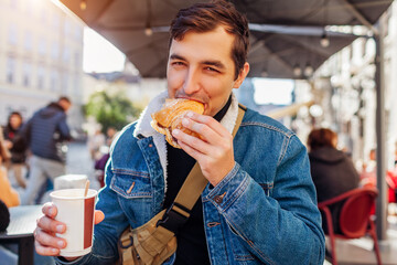 Young happy man having breakfast with coffee and croissant sitting at outdoor cafe in European city. - obrazy, fototapety, plakaty