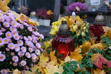 Fototapeta premium Grave with glass candles and flowers in pots among dry autumn leaves in cemetery on November day