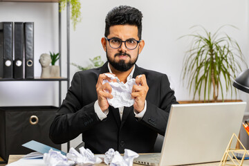 Angry furious Arab man working at office throwing crumpled paper, having nervous breakdown at work,...