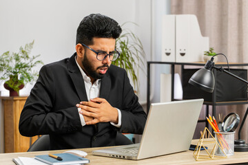 Stressed Indian businessman freelancer suffers from tension holding chest with heart pain attack risk of stroke symptom while working at modern home office workplace desk. Male Hindu in formal suit