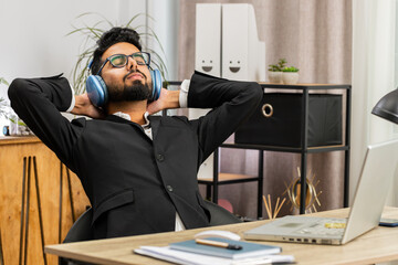 Happy smiling Indian businessman listening music on headphones taking break relaxing leaning on chair after working at office workplace. Male Hindu freelancer in formal suit puts hands behind head