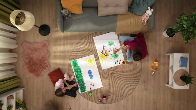 In the picture from above sit on the floor of a woman and the child they draw on the sheets of each their own. There is a sofa, on the floor are paints and toys. Theyre having fun, wondering