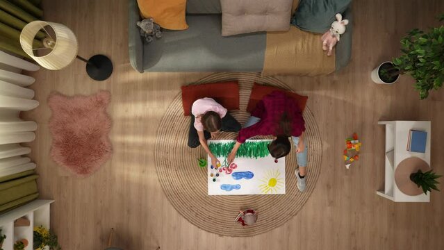 In the picture from above sit on the floor a woman and a child they together draw on paper. They communicate, spend time together. Theres a couch with toys next to it, paint on the floor. Top view