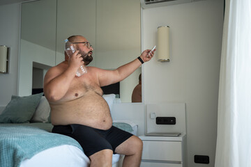 Naked obese man with bottle of cold water sweats lot adjusts air conditioner with remote control...