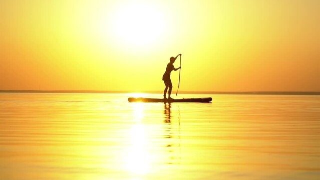 Slow motion shot of a female figure swimming on a sup puddle board into the bright orange setting sun in summer