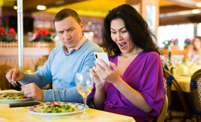 Jealous man peeking into wife smartphone, reading her messages while they have dinner in restaurant. Woman ignoring husband. Family phubbing concept..