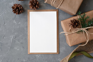 Blank paper card and crafted envelope mockup, brown gift boxes wrapped in crafted paper, decorated with pine cones and branches on gray table background. Christmas holiday greeting postcard template