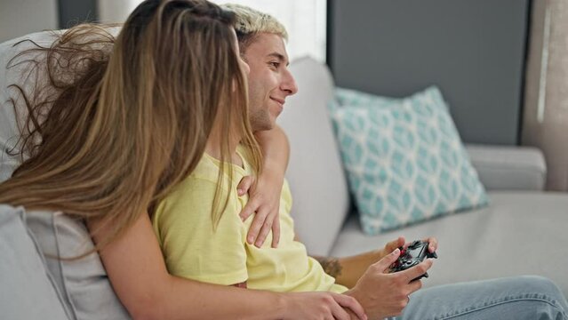 Beautiful couple playing video game sitting on sofa hugging each other at home