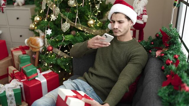 Young hispanic man make photo to gift by smartphone sitting on sofa by christmas tree at home