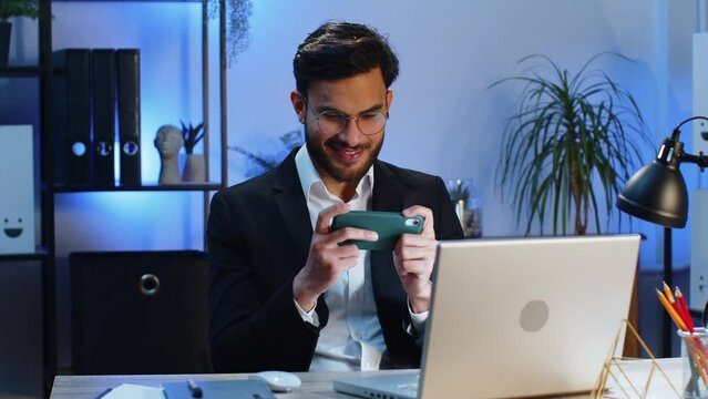 Indian businessman playing on smartphone mobile application celebrate winning racing or shooter game at office desk workplace. Professional manager freelancer man enjoy testing good work of new app