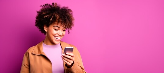 Young woman with afro hair smiling and watching her phone standing against a pink background. copy space for text