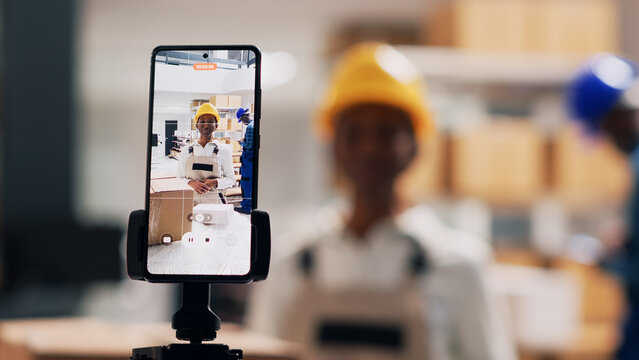 Female employee in overalls recording online video for social media app, creating advertisement for storage room products in packages. Young adult filming video on mobile phone. Handheld shot. - Powered by Adobe
