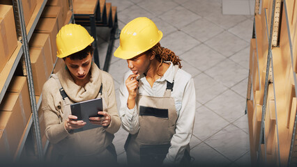 Diverse women planning order shipment with tablet, looking at cardboard packages on storage room racks. Supervisor and employee checking merchandise and products. Handheld shot.