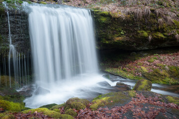 Fototapeta na wymiar Beautiful long exposure blurred motion waterfall, dark, wet cliff and red Autumn colored leaves