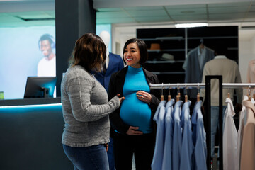 Shopping center assistant helping pregnant woman to choose casual apparel. Smiling young asian customer talking with boutique employee while searching for maternity clothes - obrazy, fototapety, plakaty