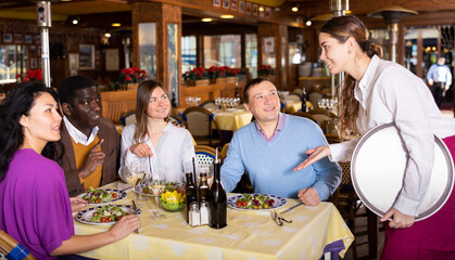 Positive company of four adults of different nationalities sitting at table in cozy restaurant during friendly meeting, talking to polite waitress