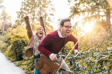 Young smiling couple riding a bicycle and enjoying fall in the city park. Attractive young people laughing and driving fast, feeling free and joyful.
