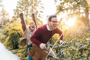 Foto op Canvas Young smiling couple riding a bicycle and enjoying fall in the city park. Attractive young people laughing and driving fast, feeling free and joyful. © Dorde