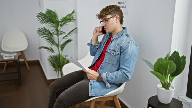 Handsome young hispanic man engaged in a digital conversation over his mobile phone, reading important paperwork while comfortably sitting on a chair within the indoors of a waiting room