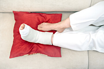 Female broken foot in white plaster cast lying on a red pillow resting. Injury, trauma, recovery,...