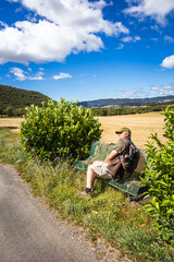 Naklejka na ściany i meble lone male hiker walking on the road, taking the opportunity to rest on a bench on the road and enjoying the sunshine