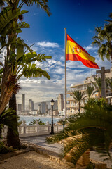 Flaga Hiszpanii na placu między palmami, a w tle widok na Hiszpańskie miasto Benidorm na Costa Blanca