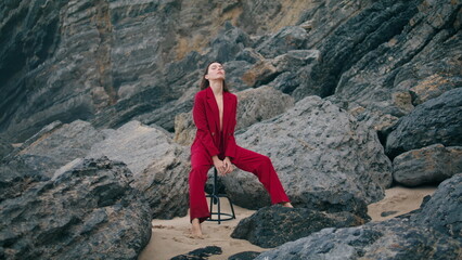 Woman posing beach cliffs wearing red suit cloudy day. Model sitting on chair.