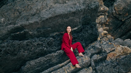 Naklejka na ściany i meble Luxury model sitting rock wearing stylish red suit. Woman posing on stone cliff