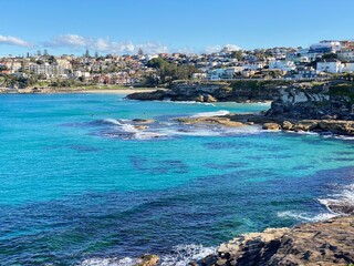 Fototapeta premium Bay and coastline next to the ocean. View of the city in the distance. View of turquoise waves breaking on the shore. Rocky coast of the ocean. Landscape and shore. Australia, Sydney.