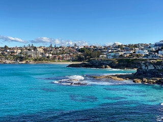 Fototapeta premium Bay and coastline next to the ocean. View of the city in the distance. View of turquoise waves breaking on the shore. Rocky coast of the ocean. Landscape and shore. Australia, Sydney.