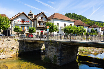 Fototapeta premium Ochagavia townscape on sunny summer day overlooking typical residential buildings with steep brown tiled roofs along stone embankment and bridge across Anduna river on sunny day, Navarra, Spain