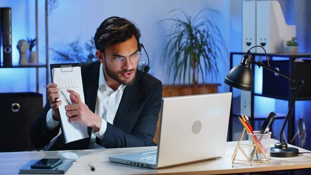 Indian businessman wearing headset, freelance worker, call center support service operator helpline, having talk with client colleague communication support showing diagram graphic statistic at office