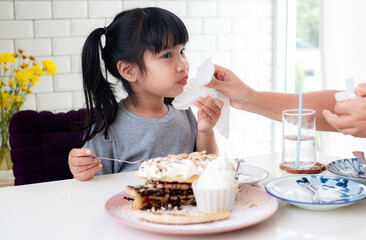 Mother's hand is using a tissue to clean her asian daughter's mouth from food stains while eating...
