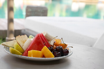 Plate with fresh fruits on table near sun lounger, space for text. Luxury resort with outdoor...