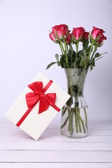 Gift box with red bow and bouquet of beautiful roses in vase on white wooden table