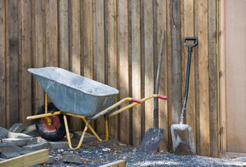 A wheelbarrow and spade placed amongst gravel and building material. Construction site at a home in the backyard. Equipment and tools used to build a house. A shovel and other building equipment - obrazy, fototapety, plakaty