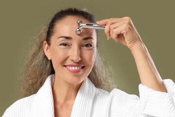 Beautiful mature woman using face massage roller on khaki background