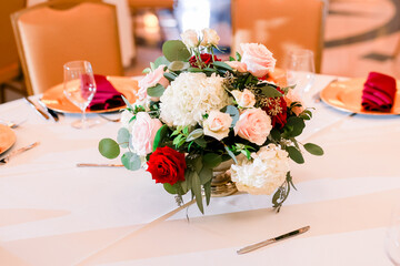 A beautiful arrangement of flowers with pink, white and red roses sits on a white tablecloth table at a wedding reception with a winter red color scheme.