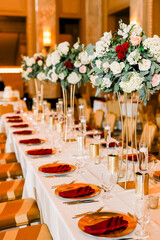 A beautiful decorated table with red napkins on gold plates and large floral arrangements, perfect for a winter wedding reception or holiday party. 