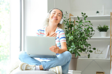 Mature woman with laptop and headphones sitting near window at home
