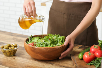 Woman adding olive oil into bowl with tasty salad at table in kitchen