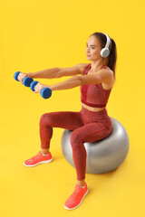 Sporty young woman with dumbbells and fitball on yellow background