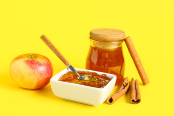 Jar and bowl of sweet apple jam with cinnamon on yellow background
