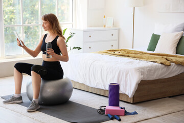 Young sporty woman with bottle of water and tablet sitting on fitness ball at home