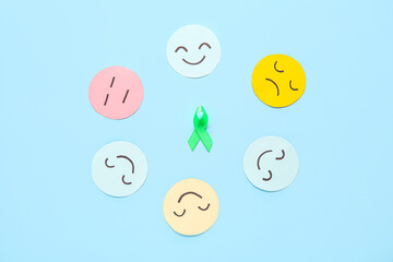 Paper faces with different emotions and green awareness ribbon on blue background. Mental Health Day