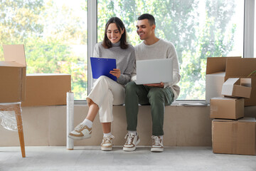 Happy young couple with laptop and clipboard in room on moving day